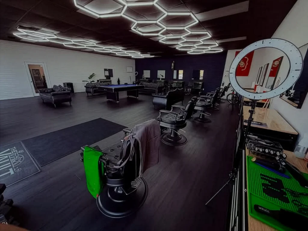 Stylish barbershop interior view with black styling chairs, hexagonal LED lighting, ring light, and pool table in the background